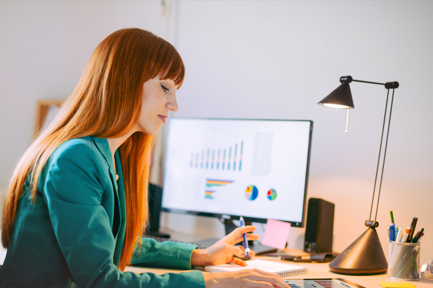 Kobieta robi notatki. W tle komputer, a na jego ekranie wykresy.