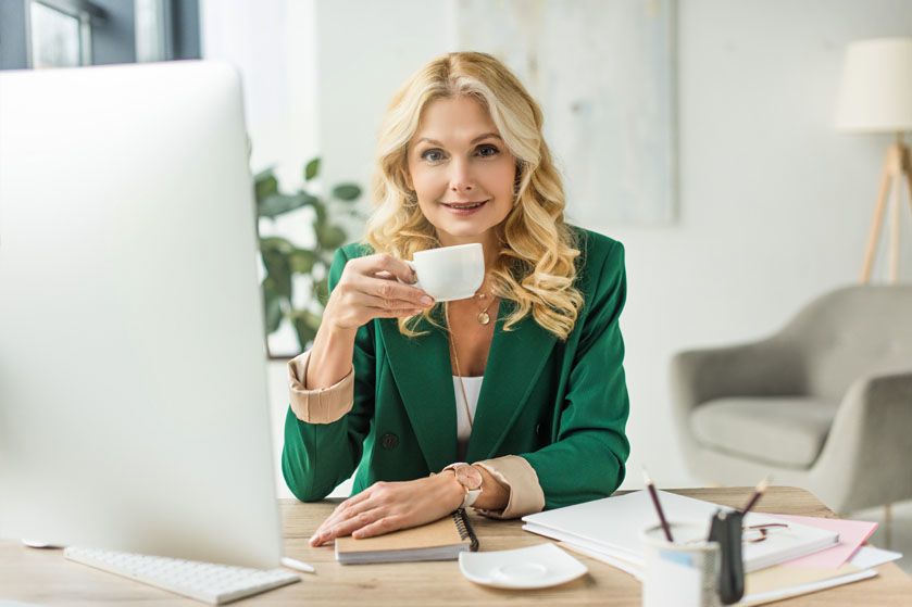 Kobieta w zielonej marynarce pije kawę przed monitorem przy biurku