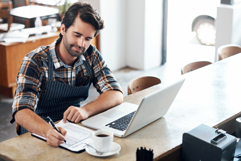 Przedsiębiorca w fartuchu sprawdza wydrukowaną listę przed komputerem i przy kawie