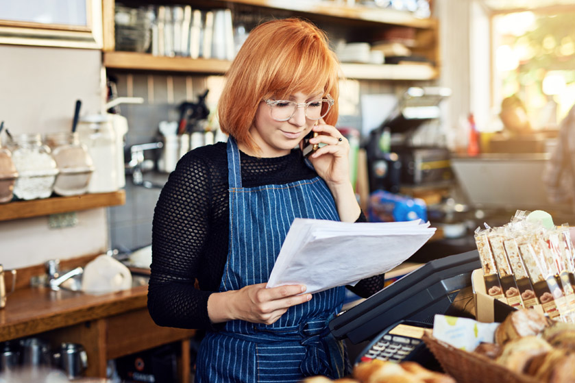 Rudowłosa kobieta w kawiarni z telefonem przy uchu przegląda plik dokumentów