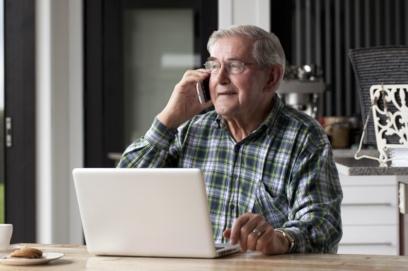 Starszy pan, który siedzi przed laptopem i rozmawia uśmiechnięty przez telefon