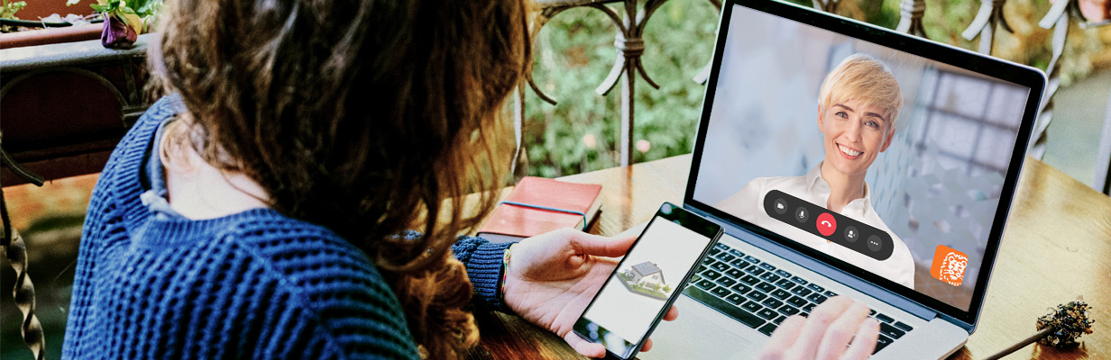 Kobieta siedząca przy biurku przed laptopem, prowadzi wideorozmowę z ekspertem hipotecznym ING. Na ekranie komputera widoczna jest twarz doradcy, a kobieta trzymając w ręku smartfon z projektem domu.