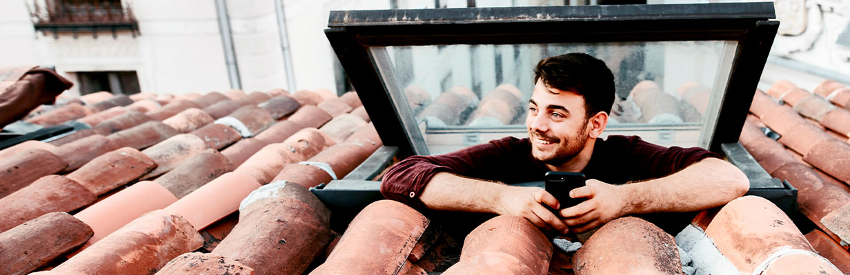 Mężczyzna wychyla się przez okno dachowe na poddaszu, uśmiecha się i trzyma w ręku smartfon, którym robi zdjęcie widoków za oknem.