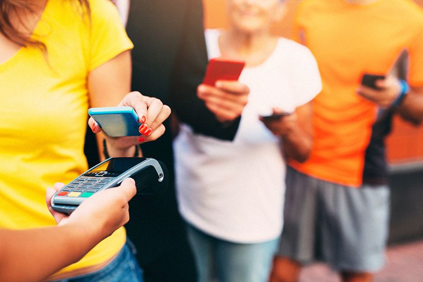 Płatność telefonem na terminalu płatniczym - zbliżenie na ręce, które trzymają urządzenia. Z tyłu kolejka ludzi ze smartfonami przygotowanymi do płatności