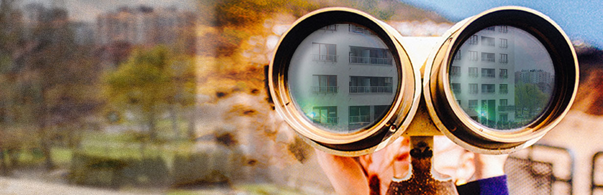 Kobieta trzyma dużą lornetkę. W szkłach odbija się oglądany przez nią apartamentowiec.