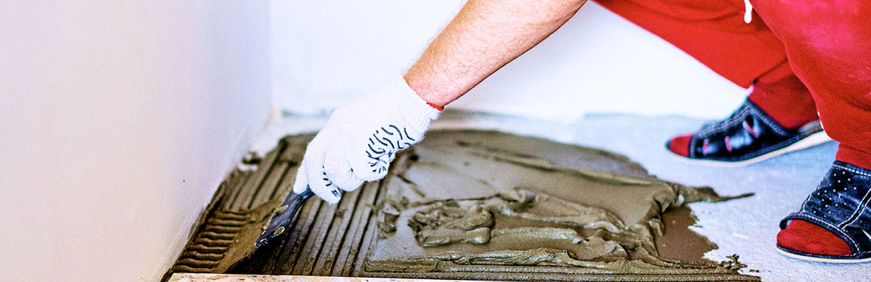 Zbliżenie na dłonie mężczyzny nakładającego klej i dopasowującego płytki podłogowe.