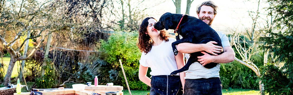 Małżeństwo w średnim wieku z uśmiechem trzyma psa na rękach w ogrodzie przy swoim nowym domu.