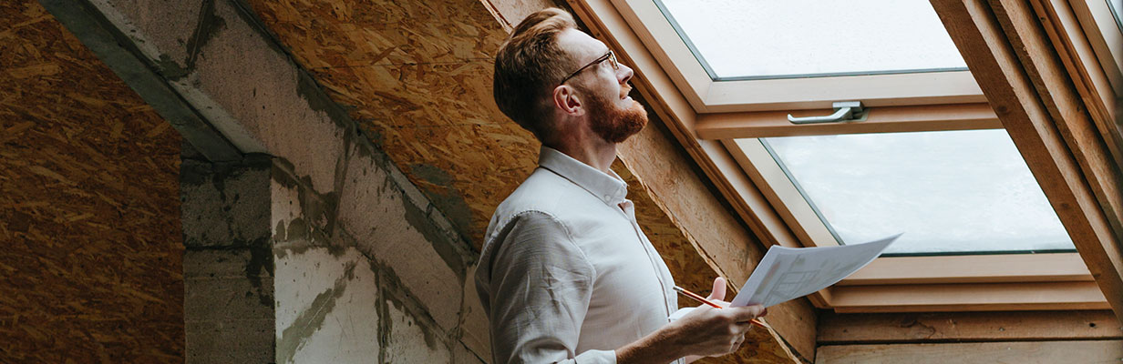 Mężczyzna stojący na poddaszu domu w stanie surowym zamkniętym, trzyma w ręku dokument – świadectwo charakterystyki energetycznej. Spogląda przez okno dachowe, przez które wpada naturalne światło, podkreślając surowe wnętrze i drewnianą konstrukcję dachu.