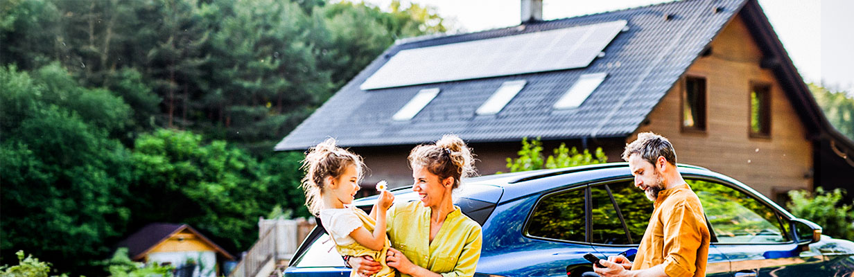 Szczęśliwa rodzina stoi przed  energooszczędnym domem z nowoczesnym dachem, na którym zamontowane są panele fotowoltaiczne.